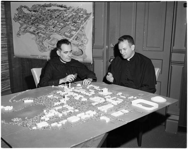 Hesburgh and Joyce sit at a table with a large 3D model of campus and its buildings. An illustrated map of campus is on the wall behind them. Father Joyce gestures at the model of the library.